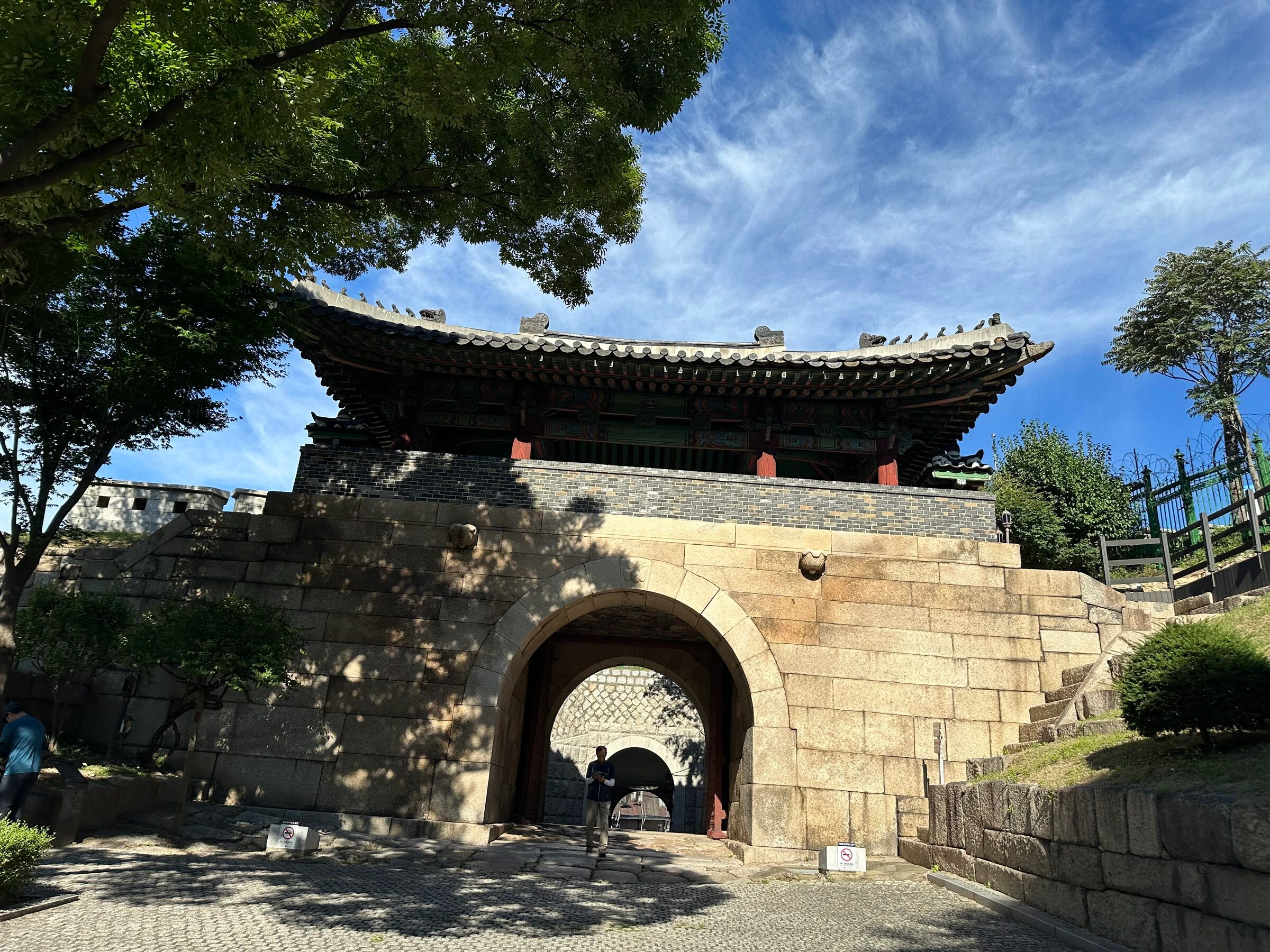 맑은 하늘 아래 창의문의 화강암 홍예문과 단청을 입힌 문루. 왼쪽에서 큰 나무가 가지를 드리우고, 그 너머 성벽 위에 원형 초소가 서 있습니다.