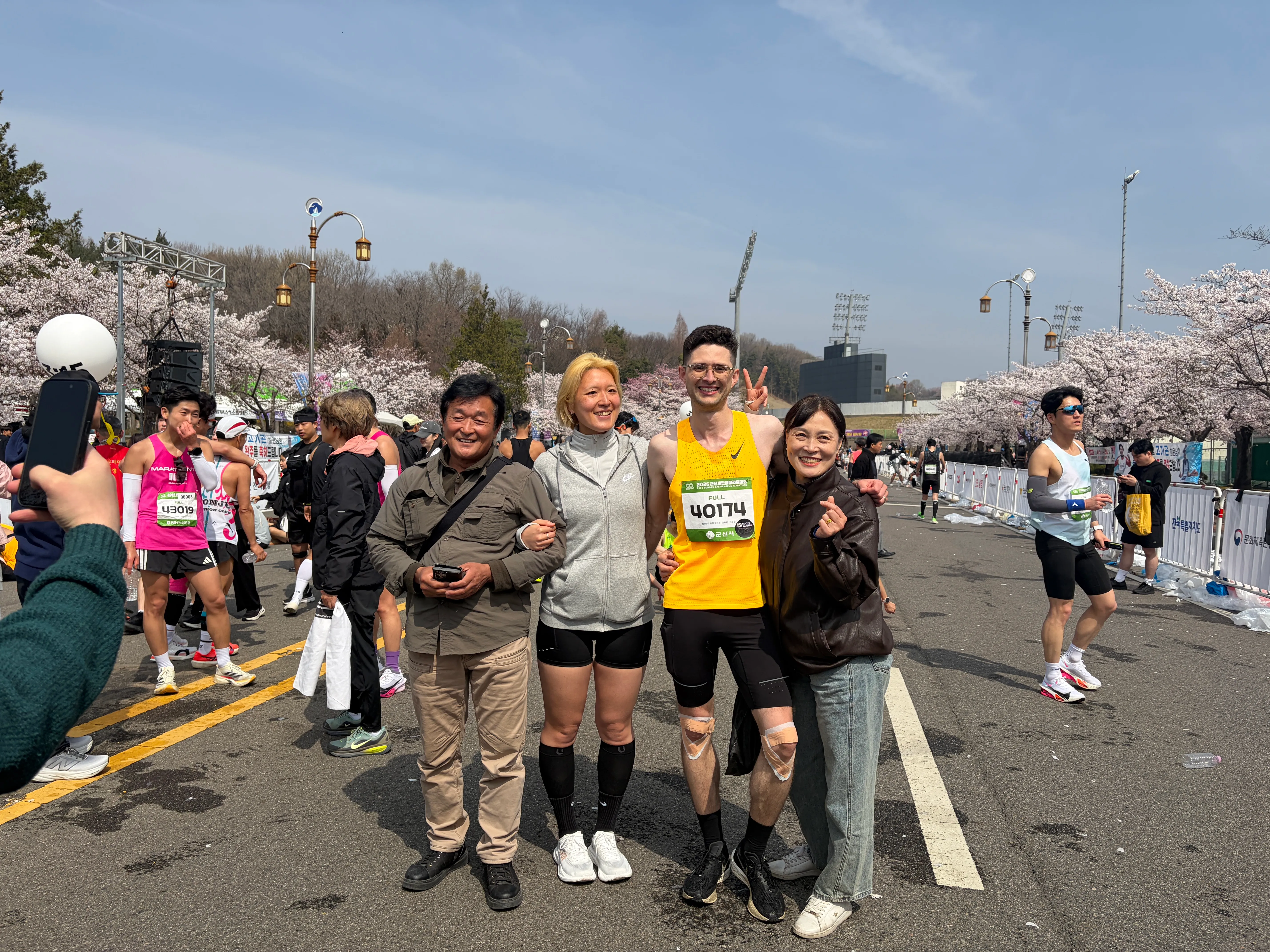 Quintin in his yellow racing kit at the 2026 Gunsan Saemangeum Marathon finish with his wife and her parents under the cherry blossoms.