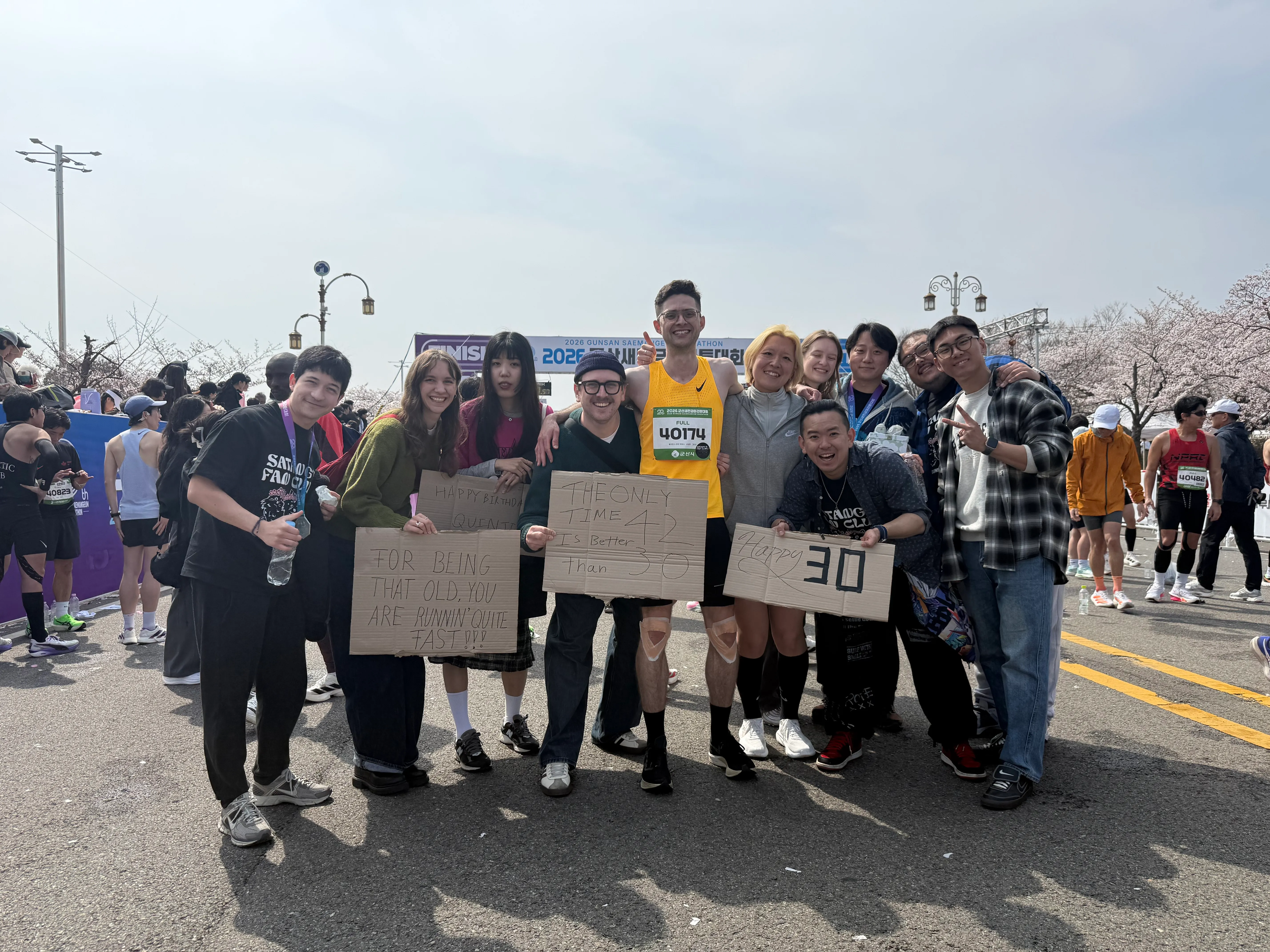 Quintin's group of friends at the Gunsan Saemangeum Marathon finish line holding hand-made cardboard signs, including one that says "30" for his 30th birthday.