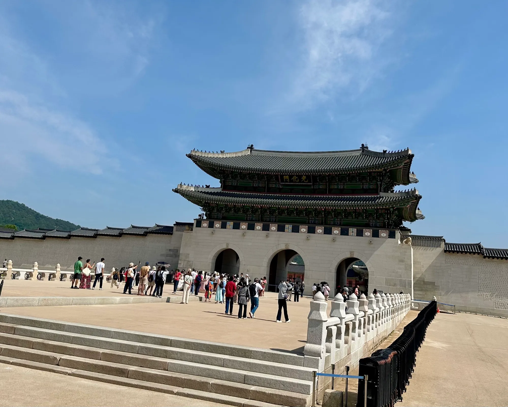 The grand two-tiered wooden gate of Gyeongbokgung palace behind its stone wall, with Bugaksan mountain to the left and visitors crossing the plaza on a clear day.