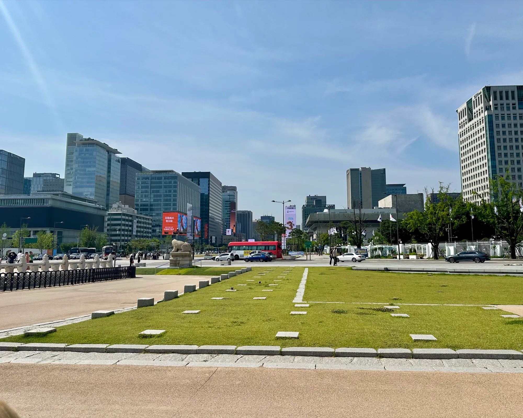Gwanghwamun Square looking south from the palace, a lawn and stone haetae statue in front of the modern Seoul skyline of skyscrapers.