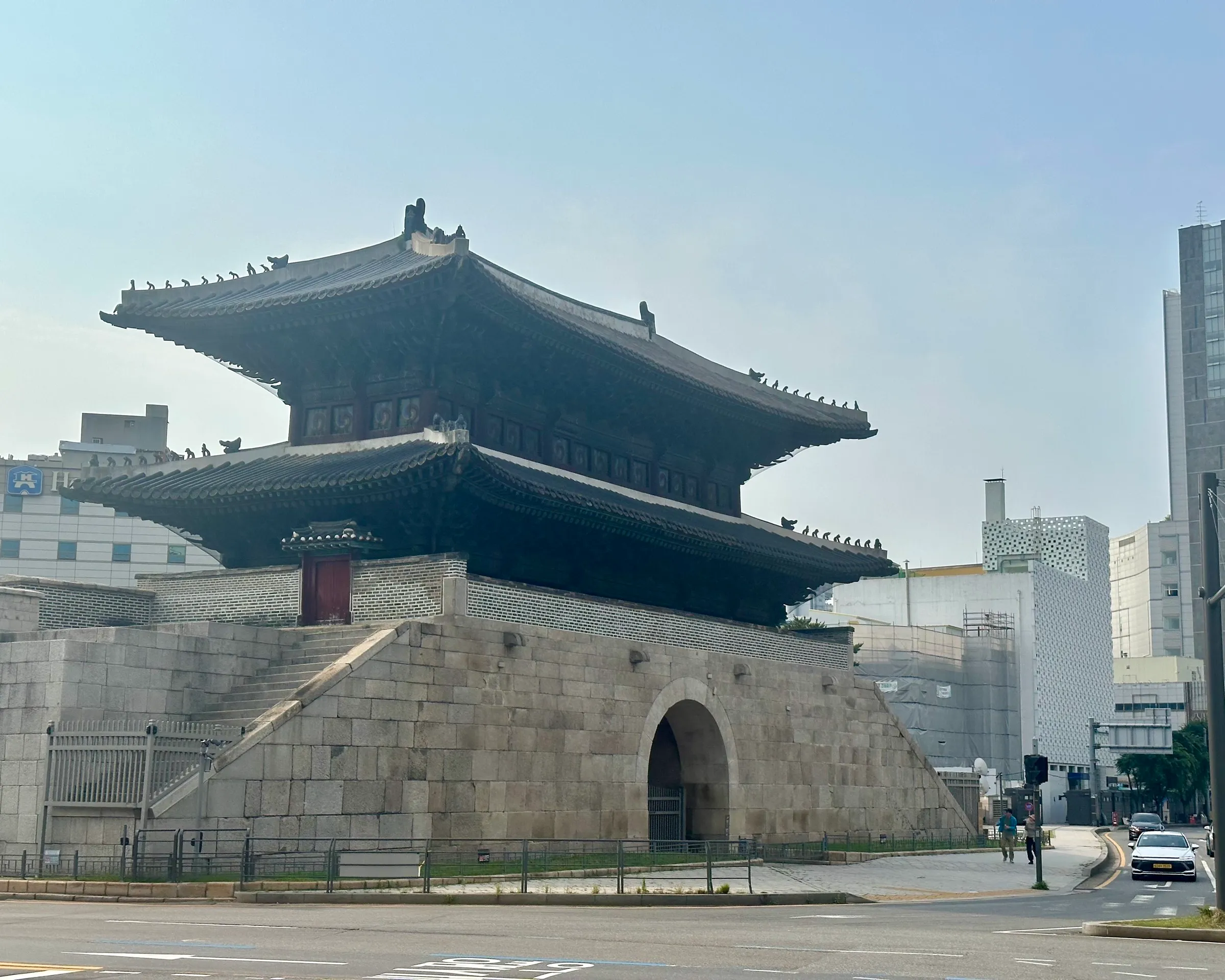 Heunginjimun, the Great East Gate (Dongdaemun), a two-tier wooden pavilion on a stone base with its arched gateway, pigeons lined along the eaves against a hazy sky.