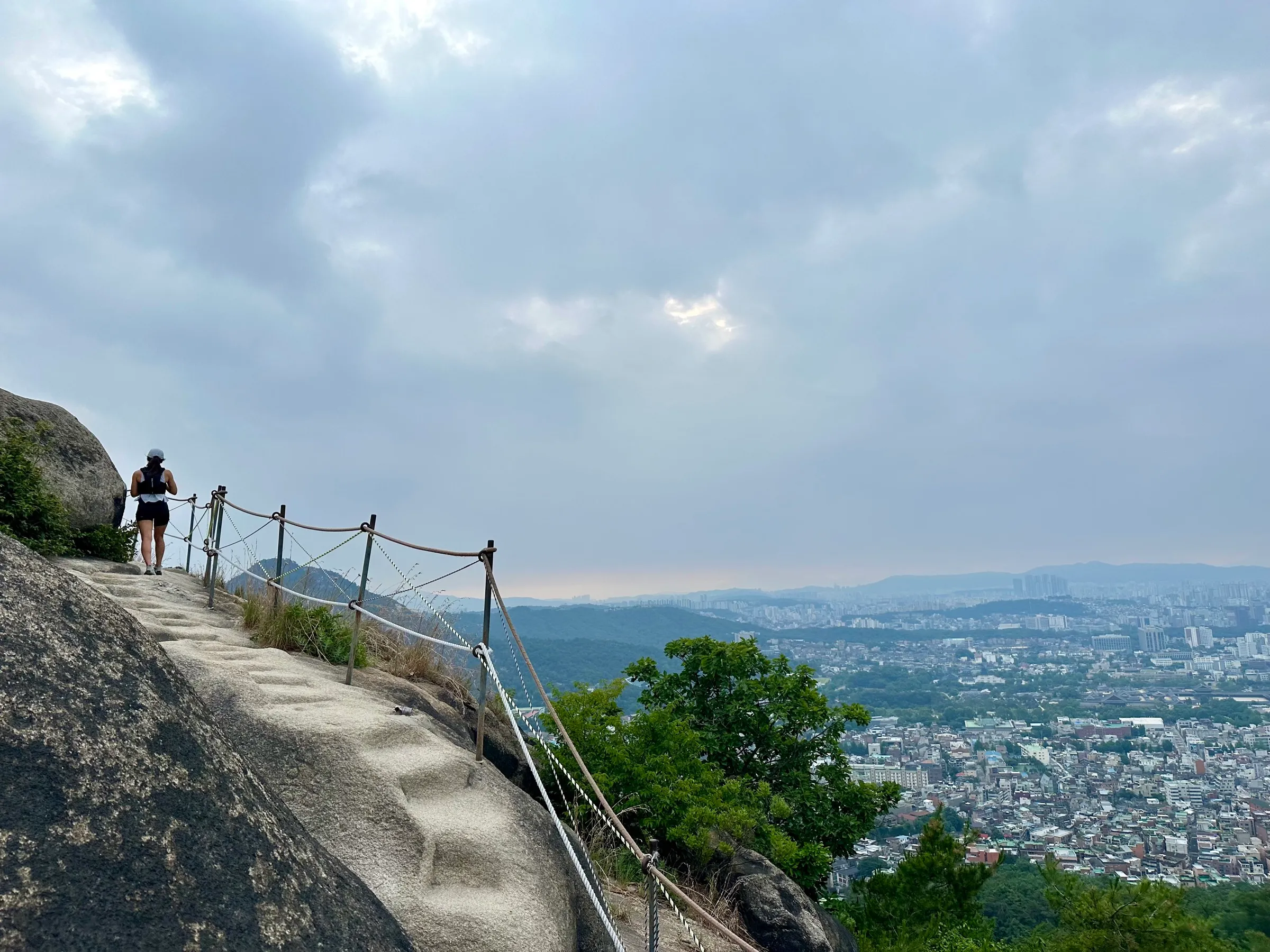 A runner in a white cap climbing the bare granite slab of Inwangsan along a rope-and-post railing, the Seoul City Wall trail, with the city spreading out far below under a moody dawn sky.