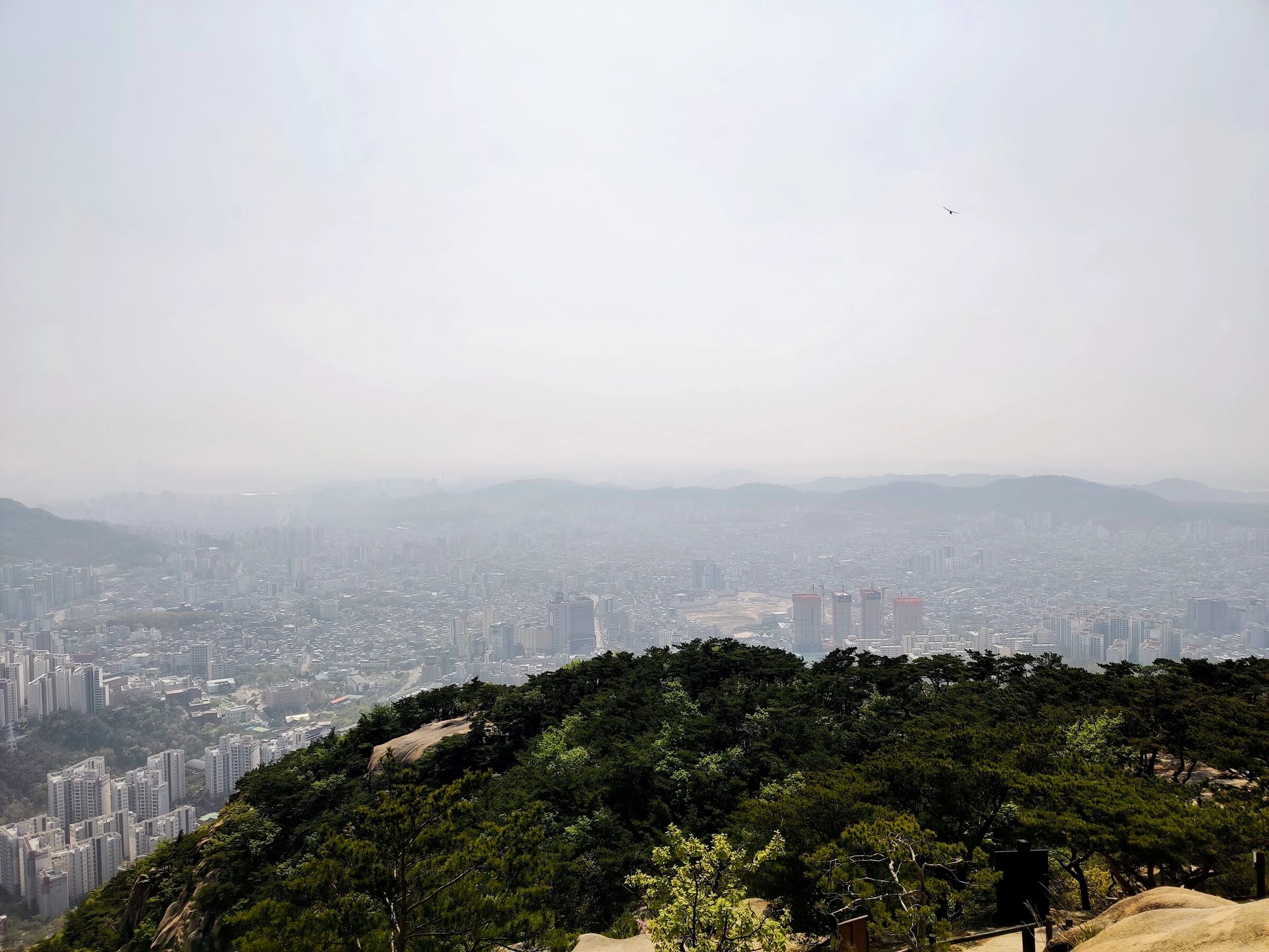 Northwestern Seoul from a granite summit on Bukhansan in April, the city flattened under spring haze with the far ridgelines barely visible.
