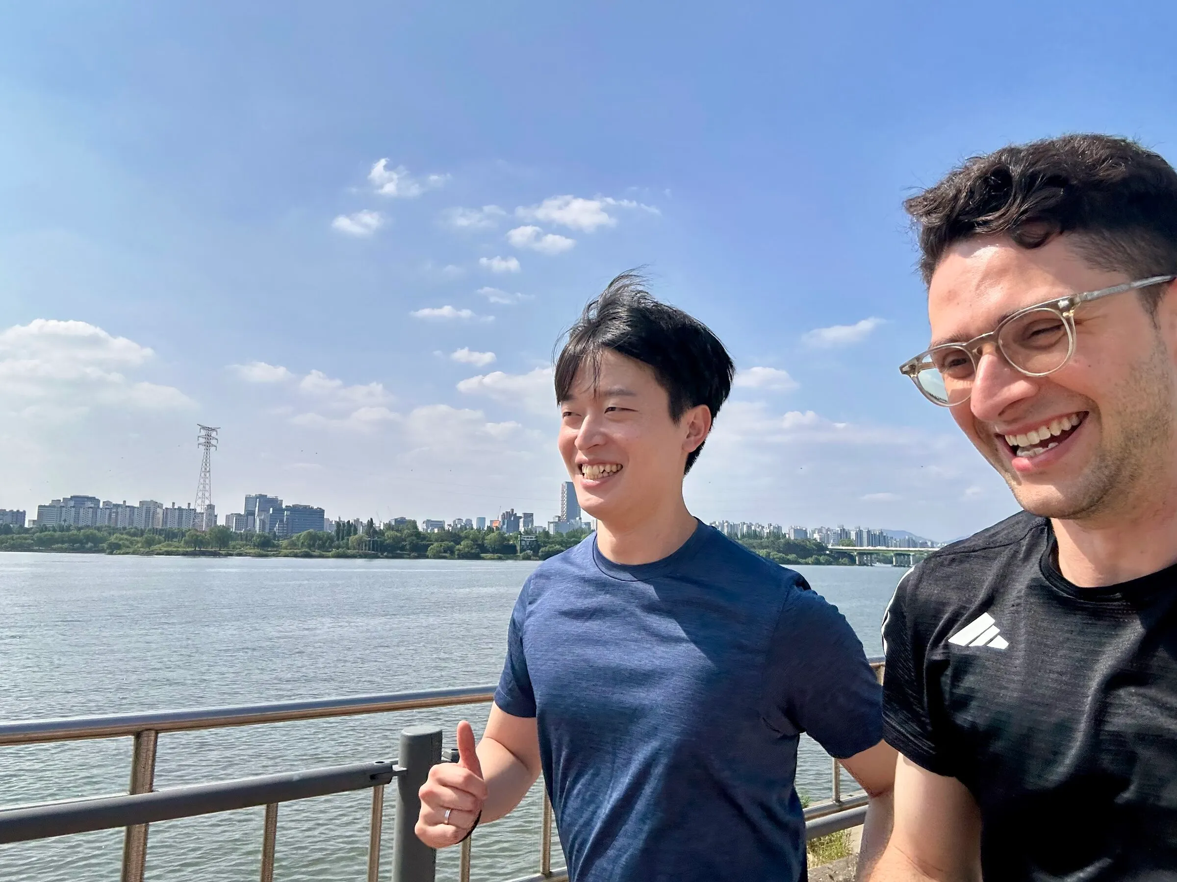 Quintin and a Run & Talk student running side by side along the Han River path, both smiling, blue sky and the Seoul skyline across the water.