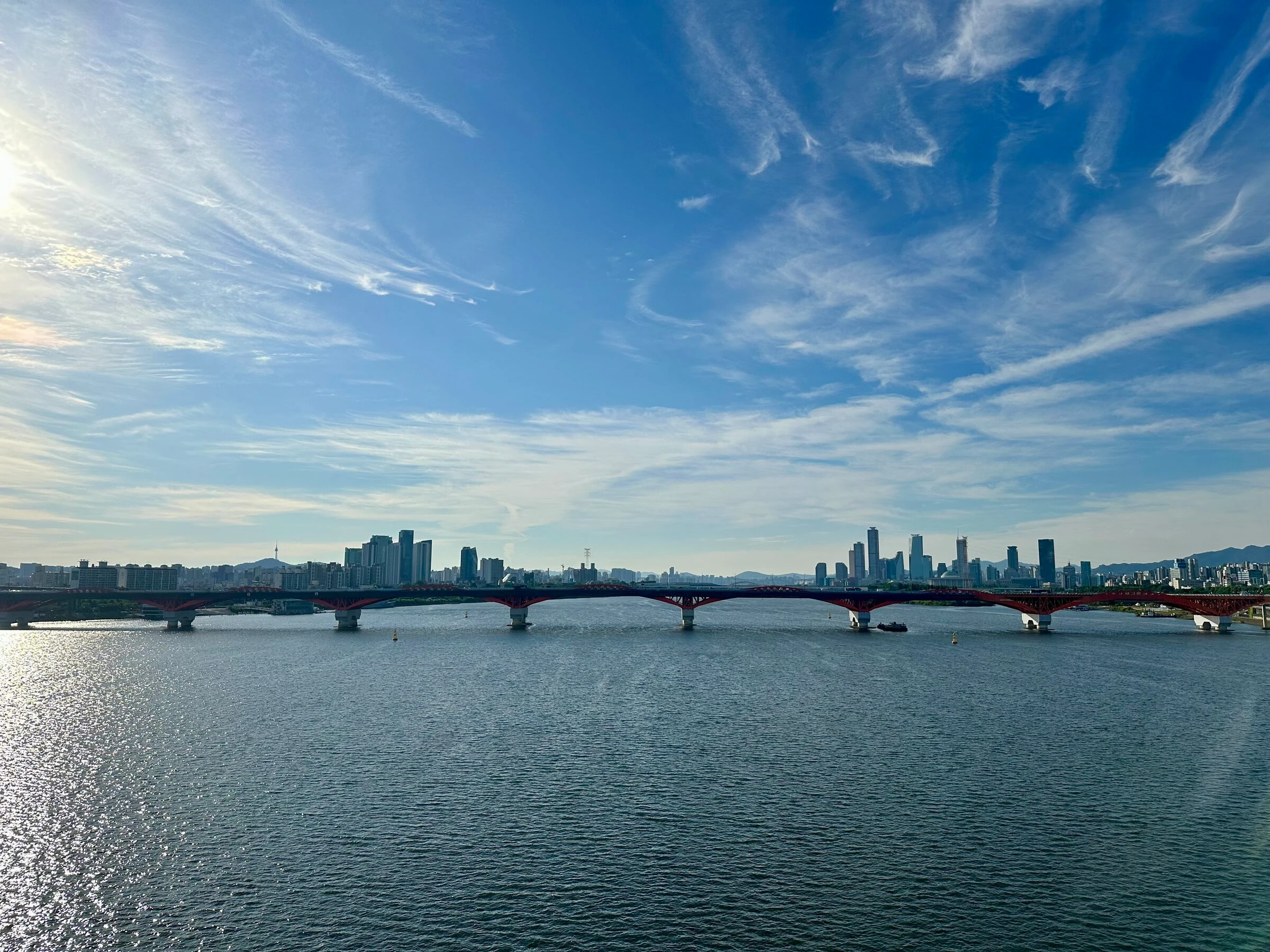 The full red span of Seongsan Bridge crossing a sunlit Han River with the Seoul skyline and Namsan Tower under sweeping cirrus clouds.