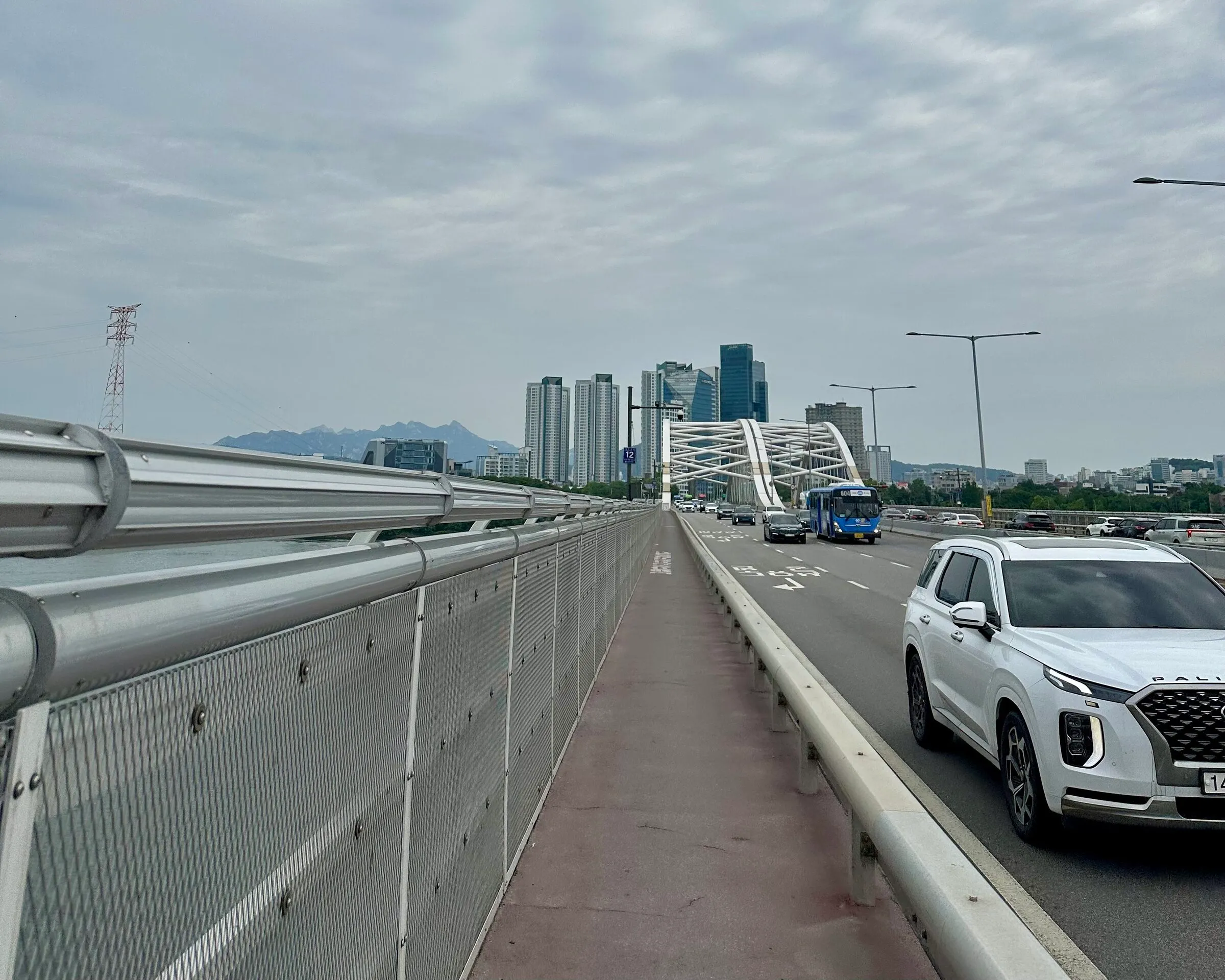 Crossing Yanghwa-daegyo on the pedestrian deck, Seoul skyline and distant mountains visible past the bridge arch.