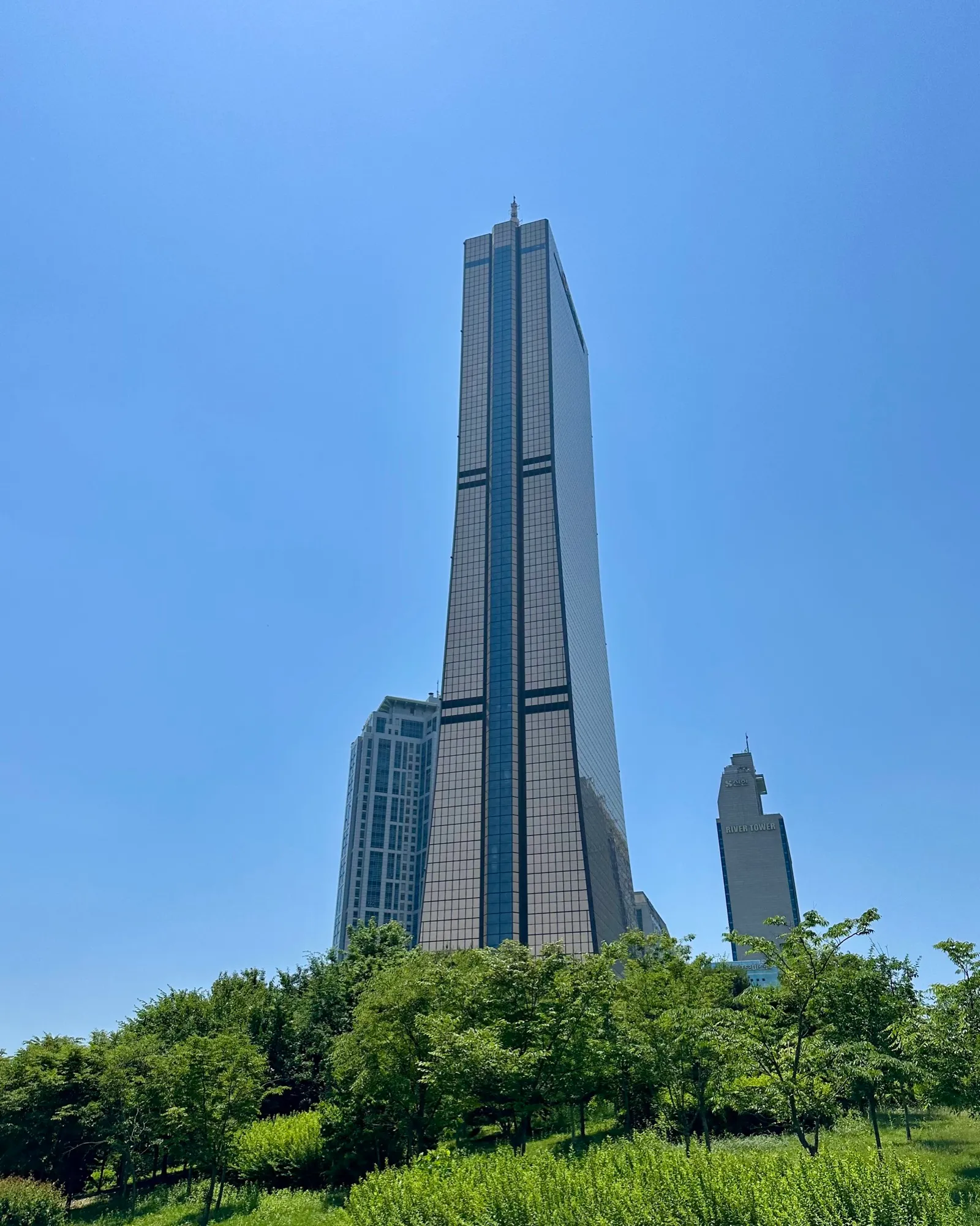 The 63 Building towering over green parkland trees against a clear blue summer sky on Yeouido.