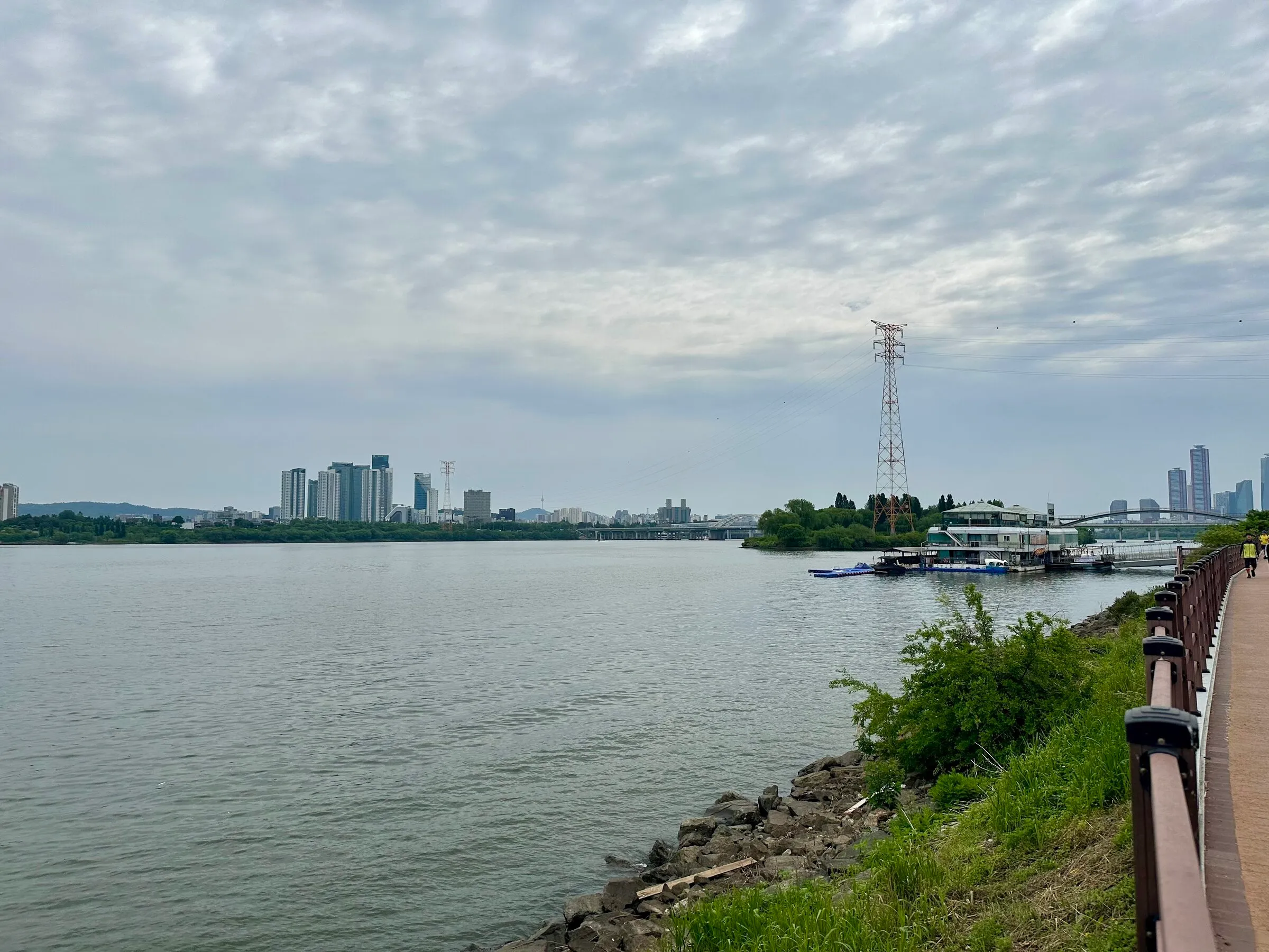 Open Han River vista from the Yeouido bank on a cloudy spring morning, the Hangang Bus floating terminal and a steel pylon on the right.