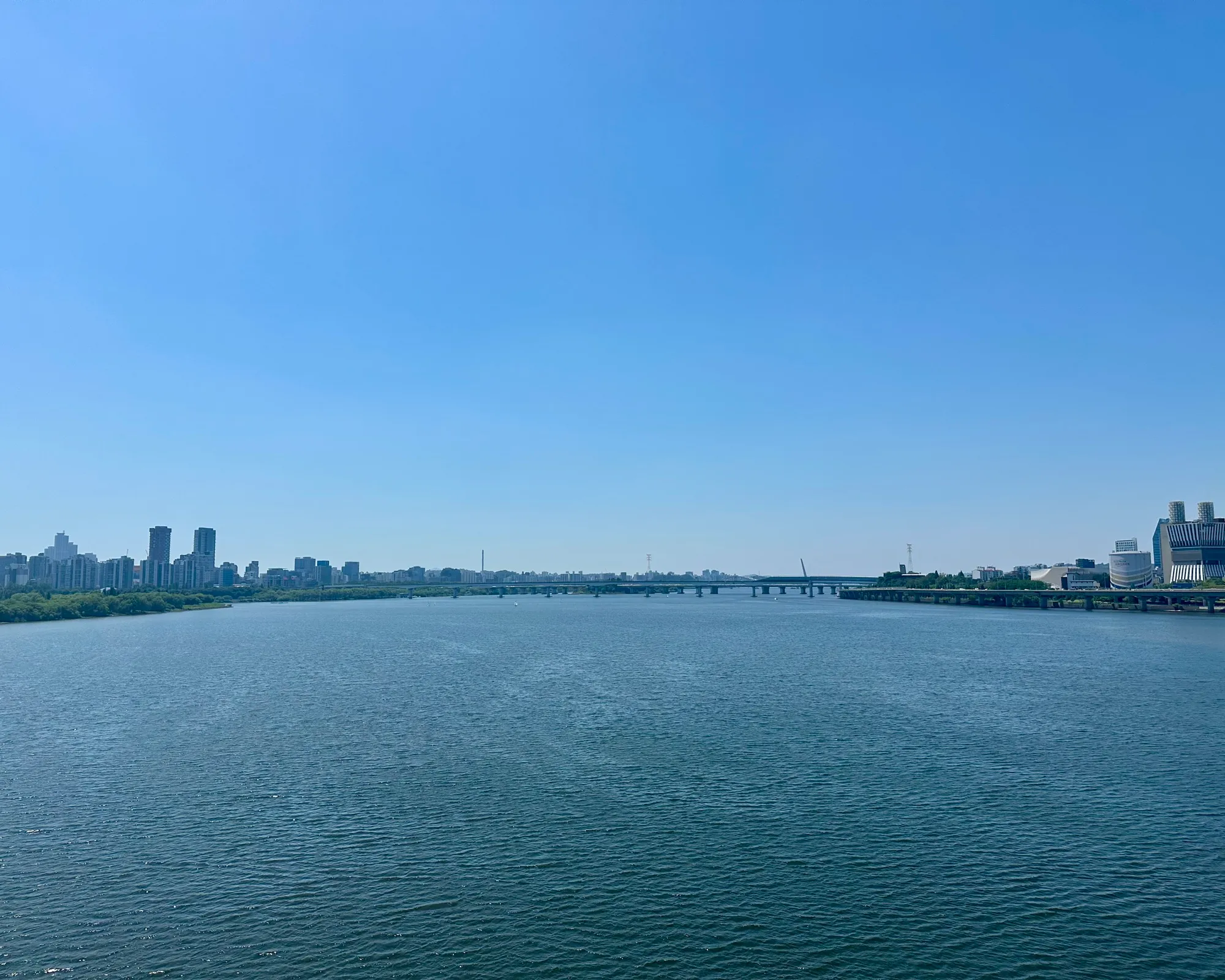 A wide open view of the Han River from a bridge on a clear blue-sky day, distant skyline and bridges along the water.