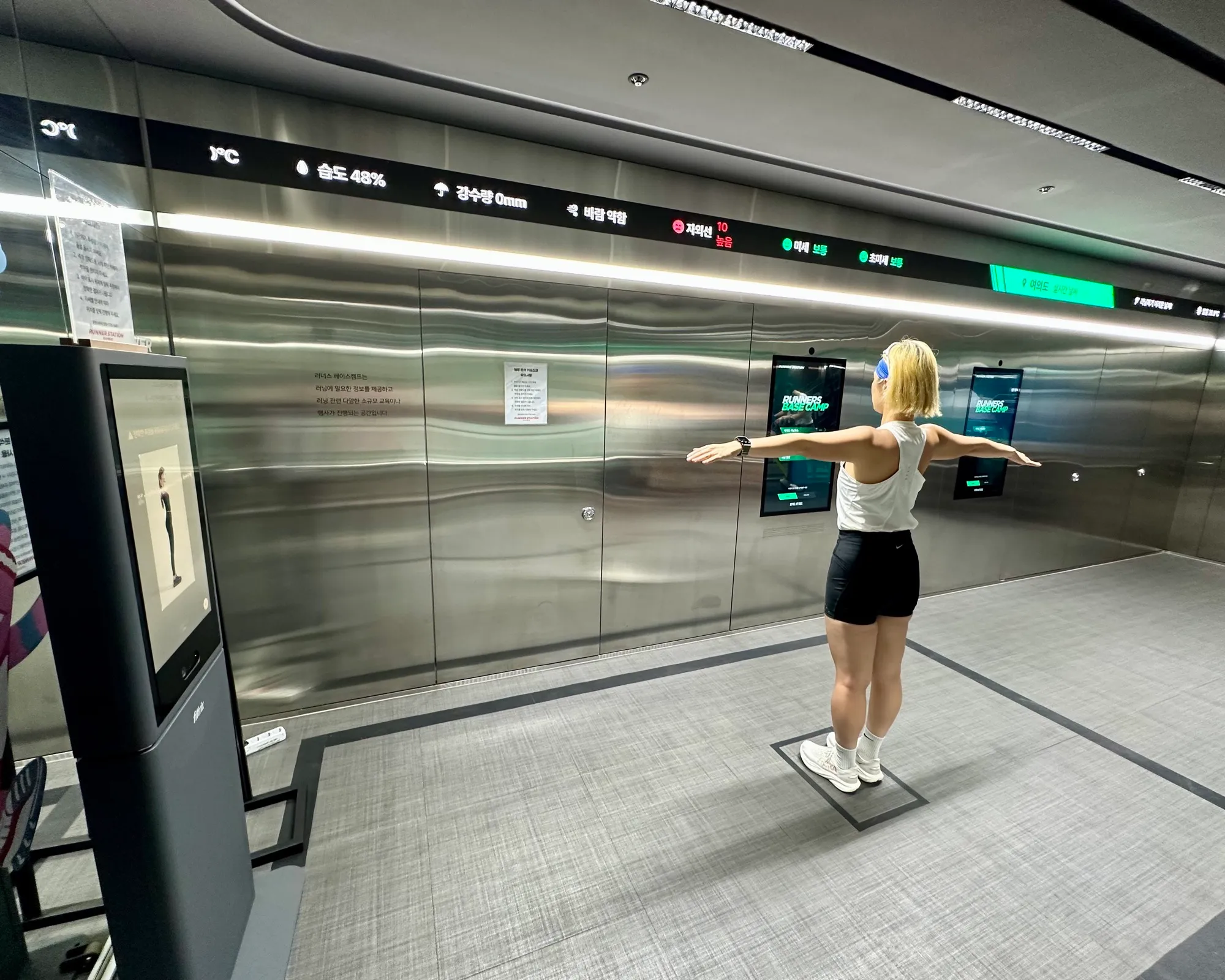 A runner stands arms-outstretched on a marked pad while the Running Station body analyzer scans her posture, a live weather and air readout running across the wall above.